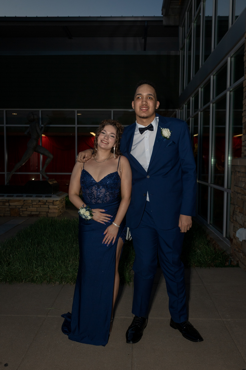 Two young people in formal blue attire pose together outside a building, with the girl in a blue lace gown and the boy in a matching suit and bow tie, smiling.