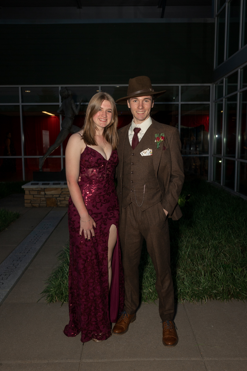 Couple in formal attire posing outdoors at night: woman in a burgundy sequined gown with a thigh-high slit, man in a brown suit and fedora.