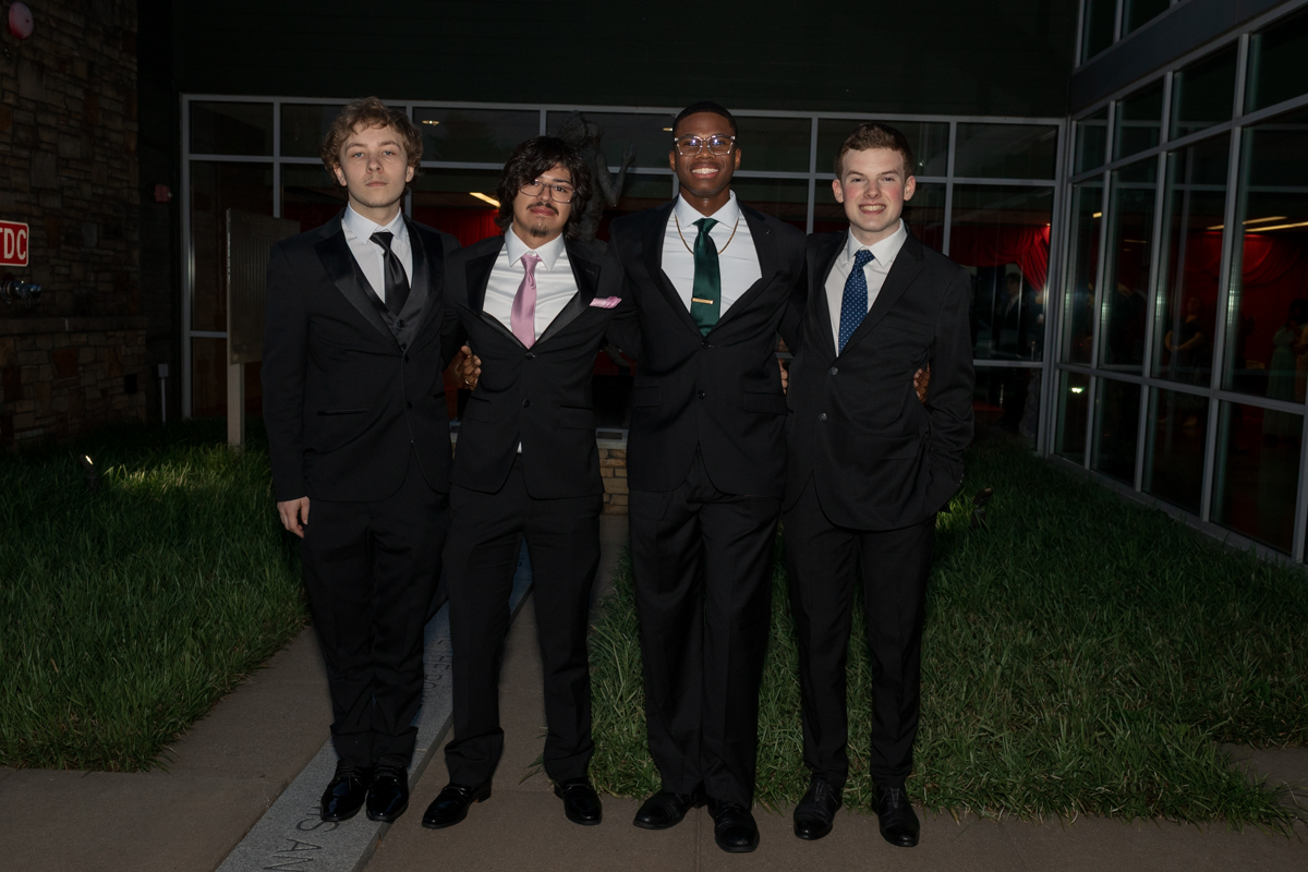 Four young men in black suits pose together outside a building, arms around each other, smiling for a group photo after a formal event.