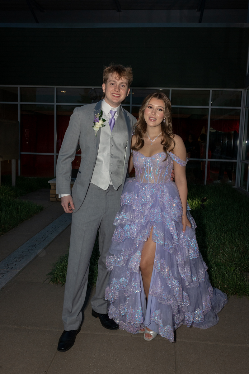 Two teens in formalwear pose together outside a building: a young man in a light gray suit with vest and lavender tie, and a young woman in a shimmering lavender ball gown with off‑the‑shoulder sleeves.