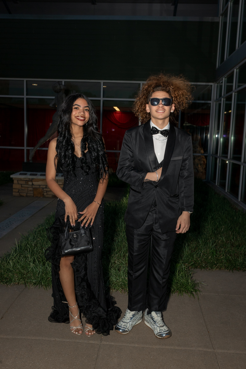 Two young people in formal attire posing outdoors at night in front of a glass building; the girl wears a black sequined gown with a high slit and handbag, the boy wears a black suit with a bow tie and sneakers, both smiling.