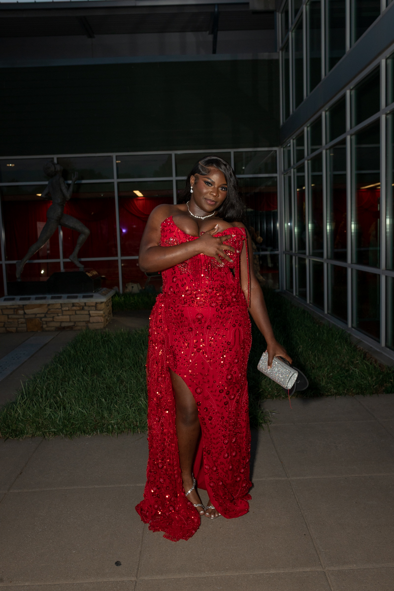 Woman in a red sequined gown posing outside a glass-front building at night, hand on her chest, holding a silver clutch.