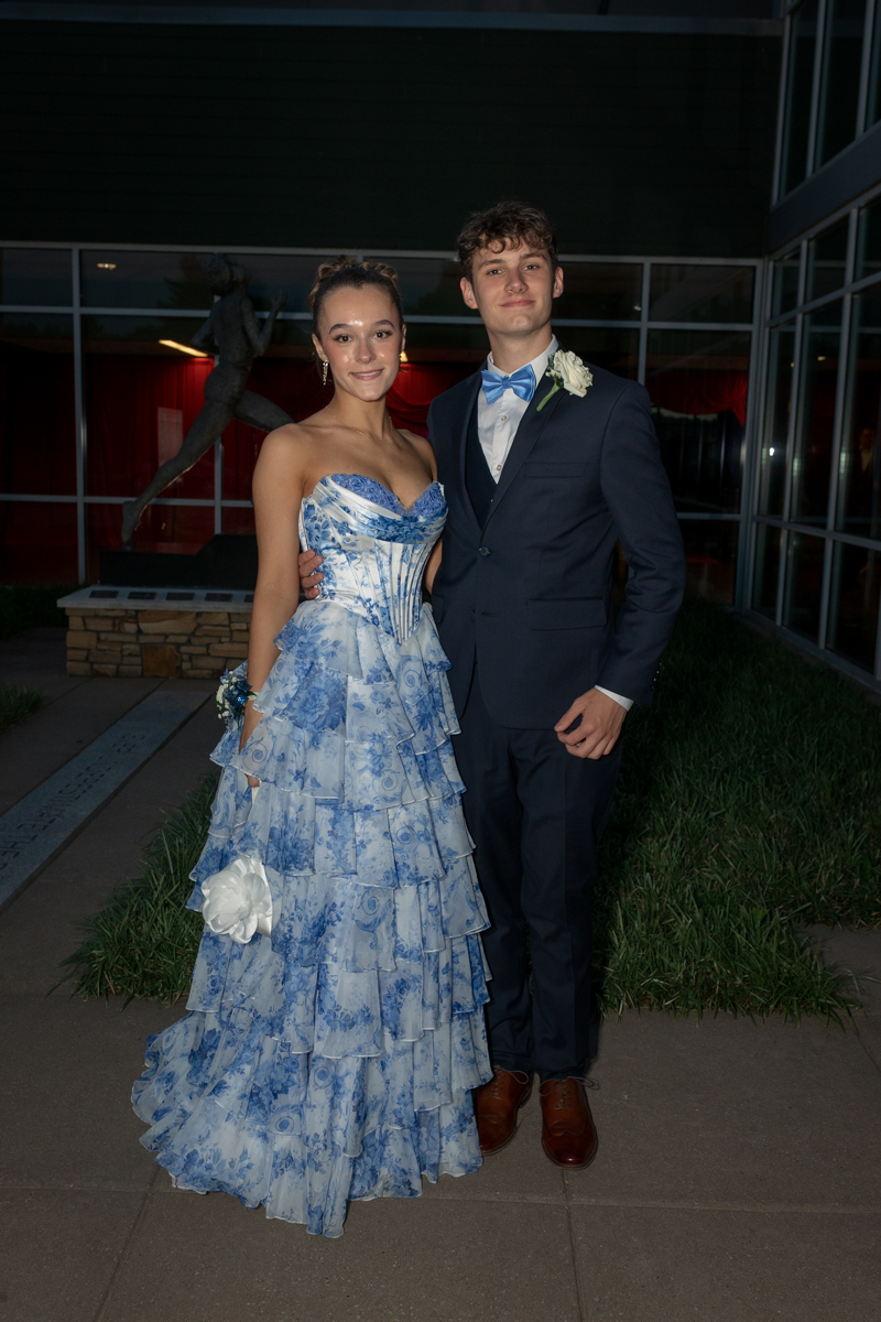 Couple in formal attire posing outside a glass-walled building; woman in a blue tiered floral gown, man in a navy suit with a light-blue bow tie and boutonniere.
