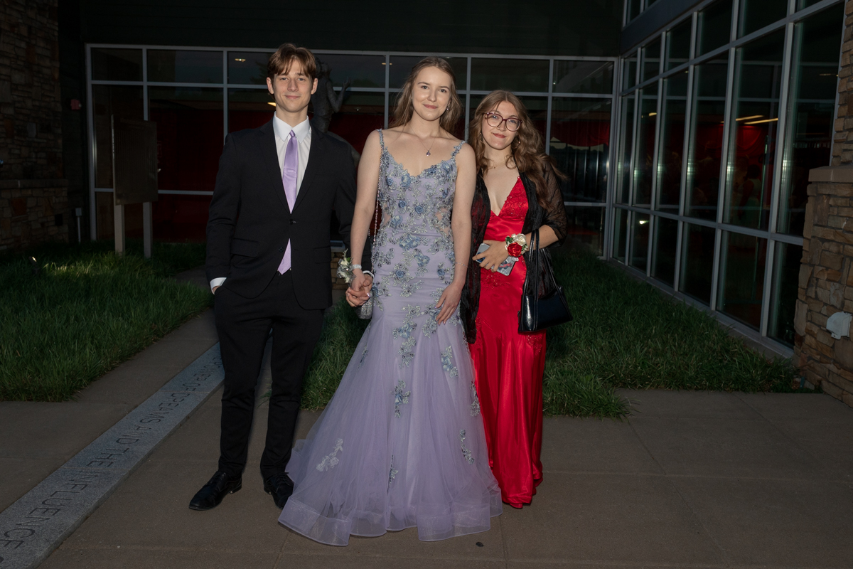 Three teens in formalwear pose outside a glass-front building: a boy in a black suit with lavender tie, and two girls in lavender floral gown and red satin dress.