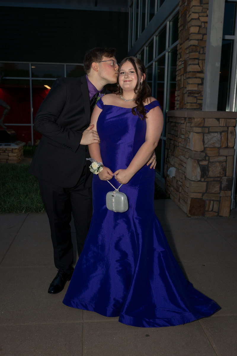Young couple in formal attire: man in a black suit kissing the woman's cheek as she poses in a royal blue gown, holding a small silver clutch