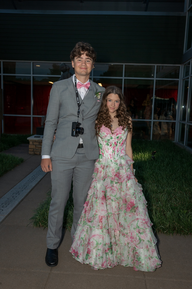 Young man in a gray suit with a pink bow tie and a camera around his neck stands next to a young woman in a pink-green floral ball gown outside a glass-front building.