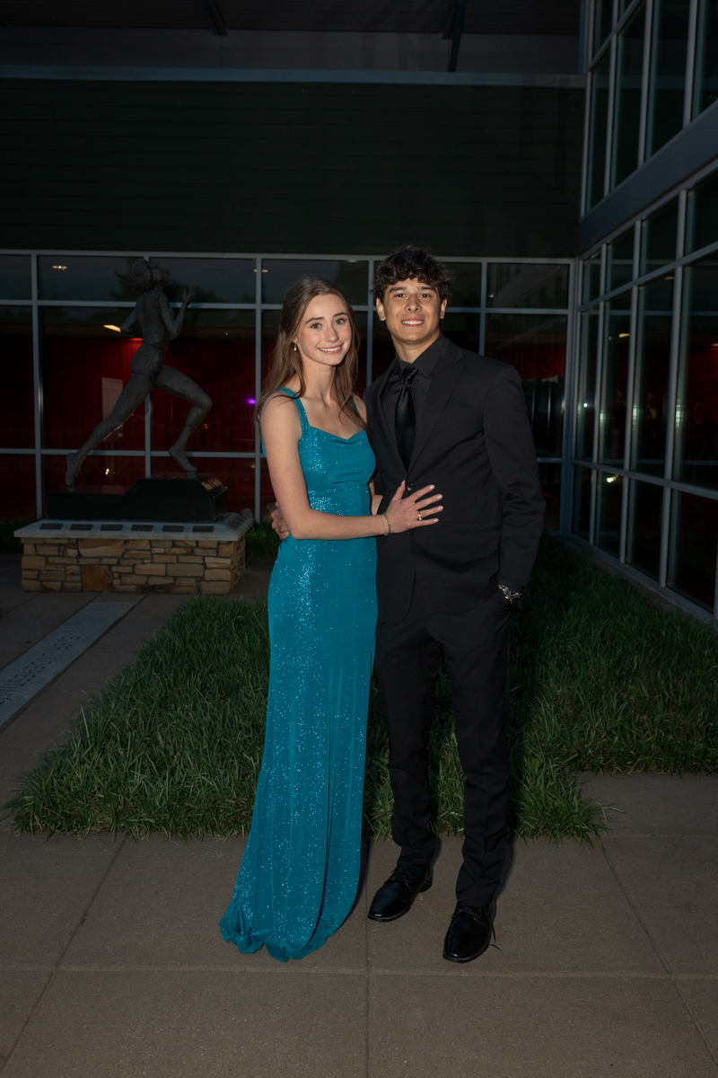 Young couple in formal attire pose together outside a glass-building venue at night, with a sculpture in the background.