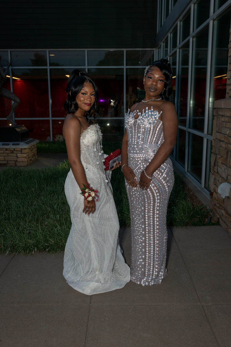 Two women in evening gowns pose outside a glass-front building; one in a white beaded strapless dress with a corsage, the other in a silver sequined gown with sheer panels.