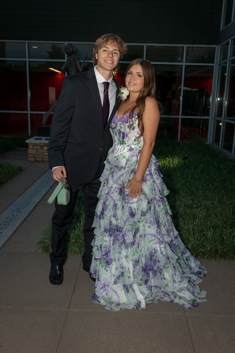 Couple posing outside a building in formal attire: man in a black suit and purple tie, woman in a lavender-green floral ball gown.