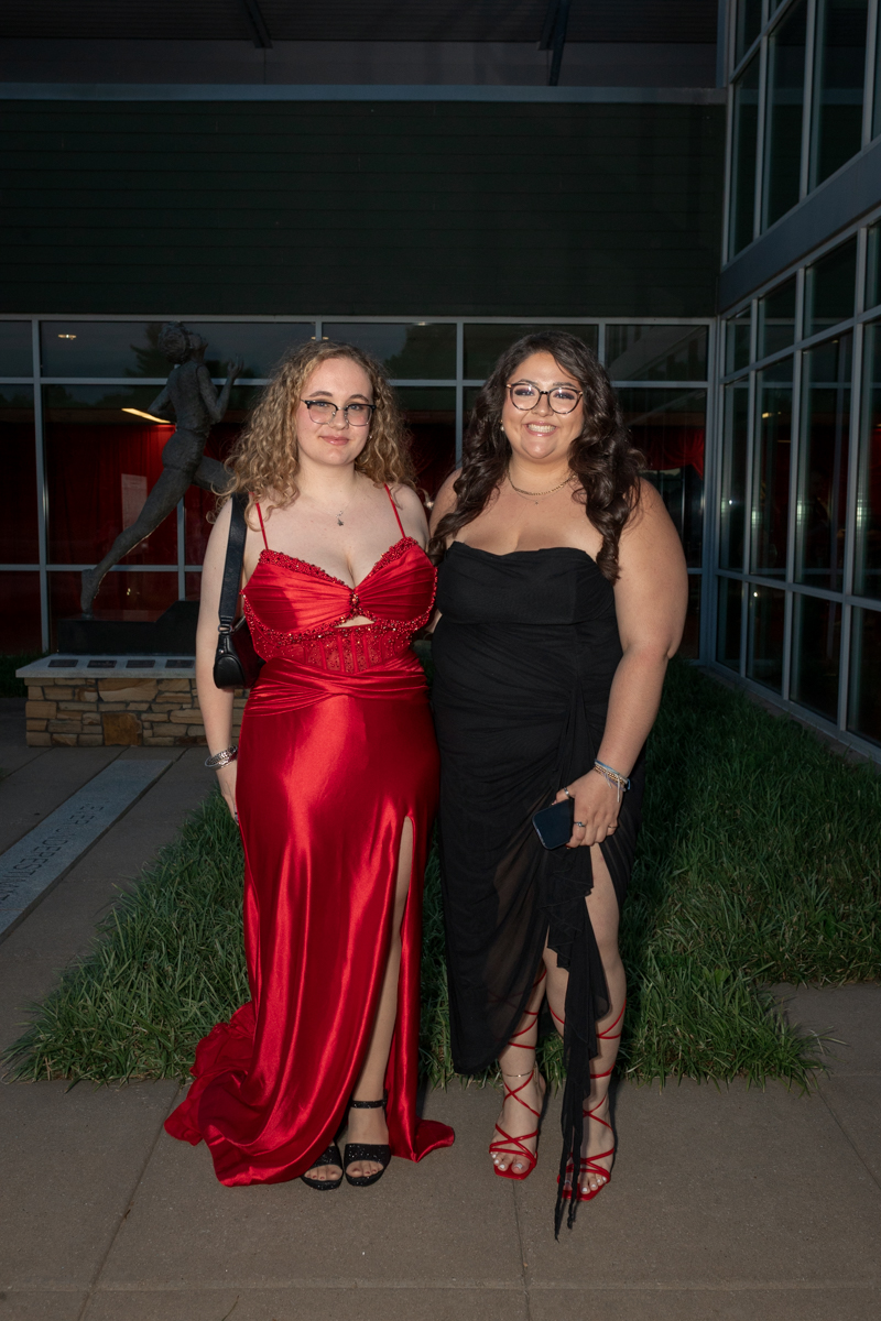 Two young women in formal dresses pose outside a glass-front building; left in a red satin gown with a thigh-high slit, right in a black gown with a wrapped skirt and red strappy heels, both smiling.