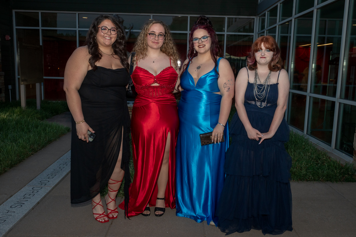 Four women in formal gowns pose outside a glass-front building, each wearing a different color dress (black, red, blue, navy).