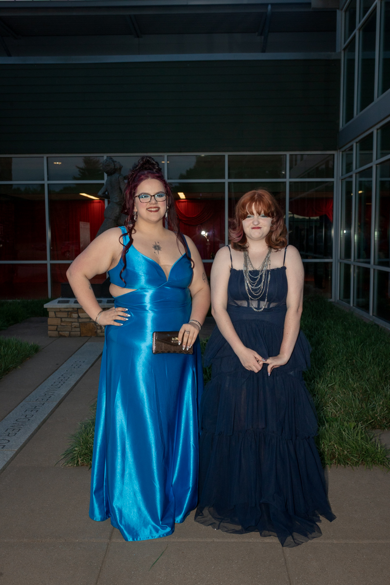 Two women in formal gowns posing outdoors in front of a glass building, smiling at the camera (one in a bright blue dress, the other in navy).