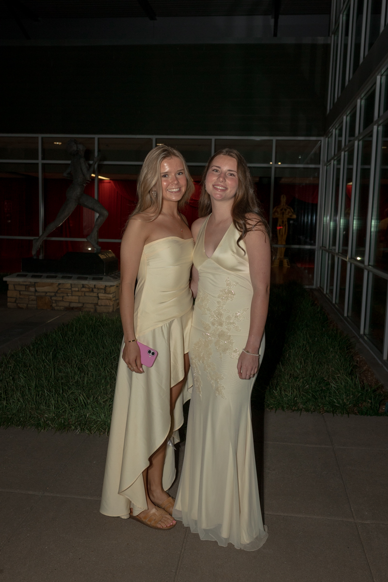 Two young women in cream formal gowns pose together outside at night, smiling for the camera nearby modern glass walls and greenery.