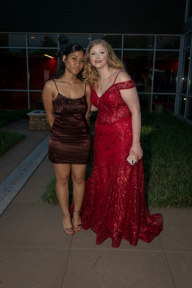 Two young women in formal dresses posing outdoors at night in front of a glass-front building with greenery nearby.