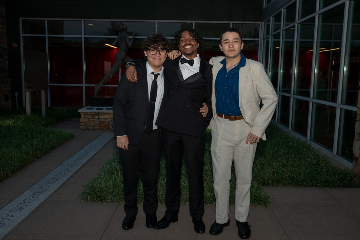Three young men in suits posing together outside a glass-front building, smiling at the camera.