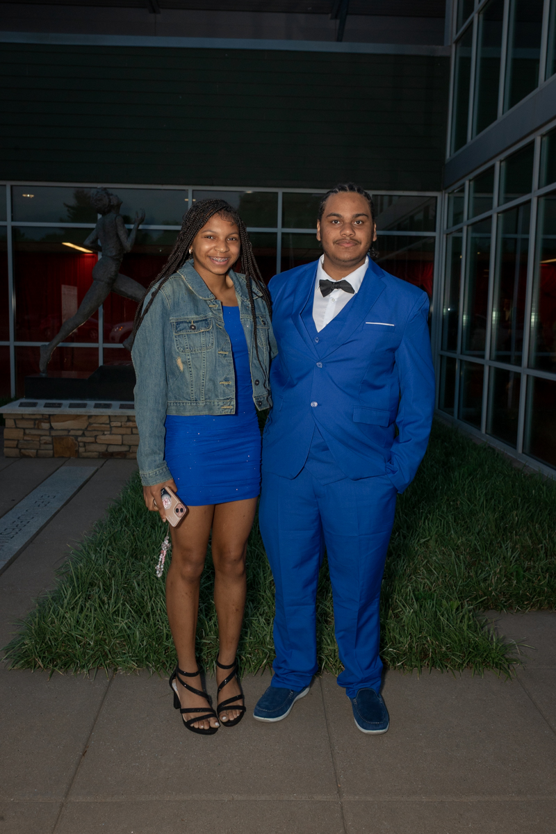 Two young adults stand side by side outdoors at night, the woman in a blue dress and denim jacket, the man in a blue suit with a bow tie.