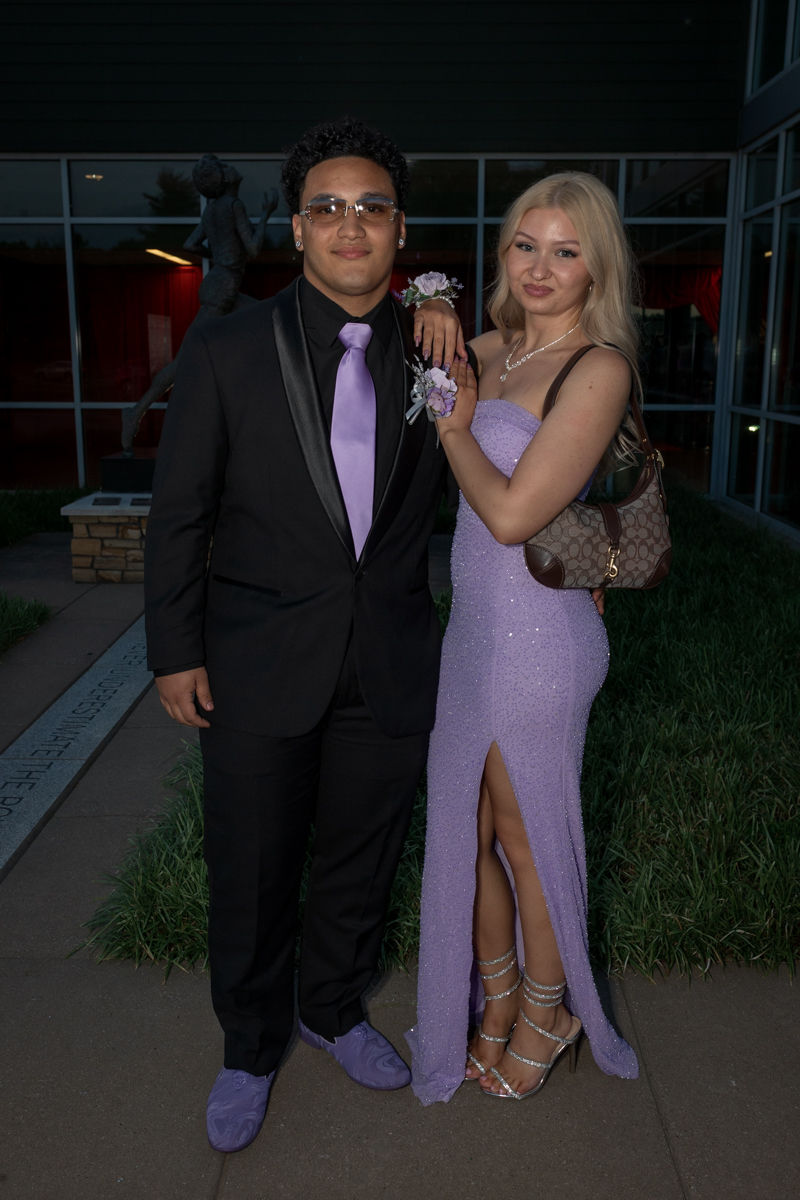 Couple posing in formal attire: man in a black suit with lavender tie and matching lavender shoes, woman in a sparkling lavender dress with high slit and silver heels.