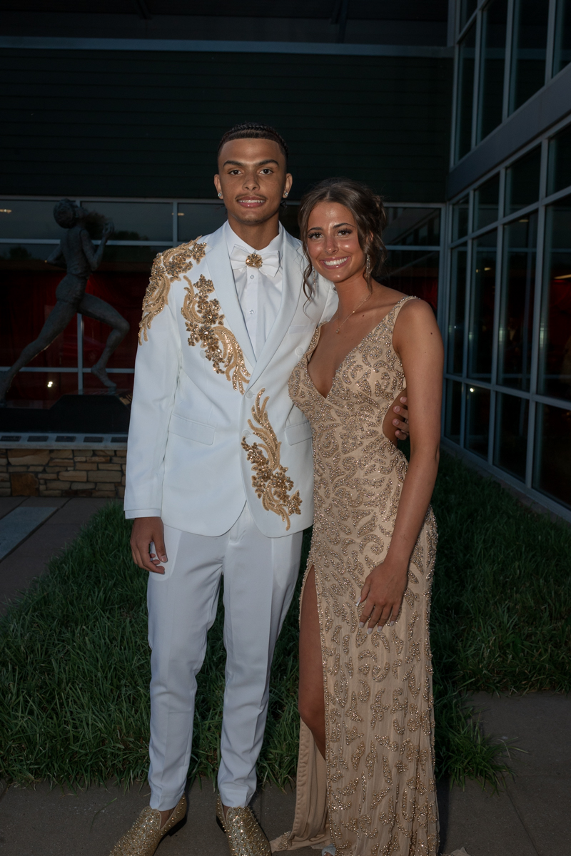 Couple posing in formal wear outside a glass building: man in a white suit with gold embroidery and bow tie, woman in a beige sequined gown with a thigh-high slit.