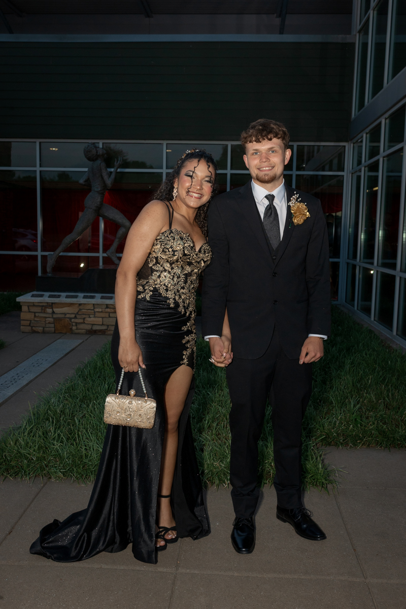 Couple dressed formally posing outdoors, holding hands; woman in a black and gold gown with a high slit, man in a black suit with boutonniere.