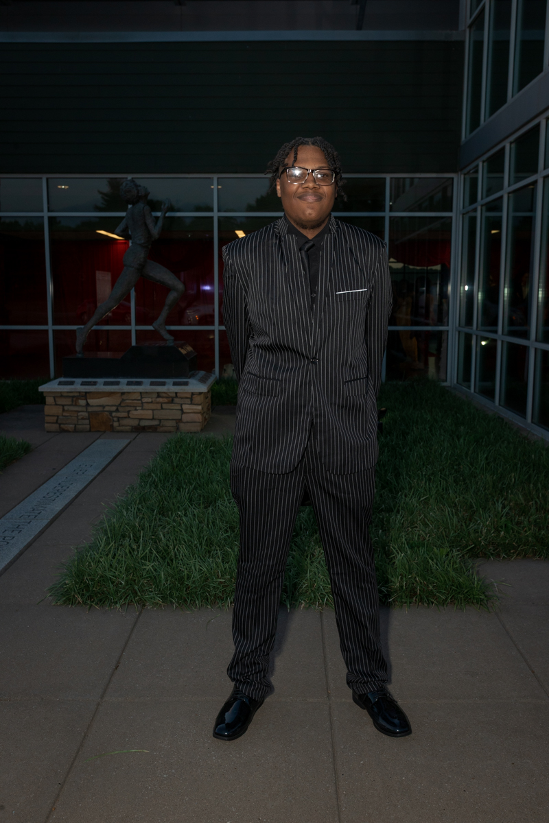 Young man in a black pinstripe suit and glasses stands outdoors in front of a glass-fronted building.