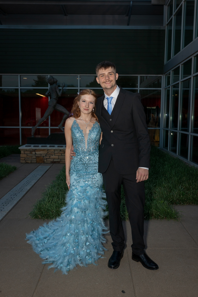 A couple posing outdoors in front of a modern glass building; the woman wears a blue sequined gown with a feathered train and the man a black suit with a blue tie, arm around each other.