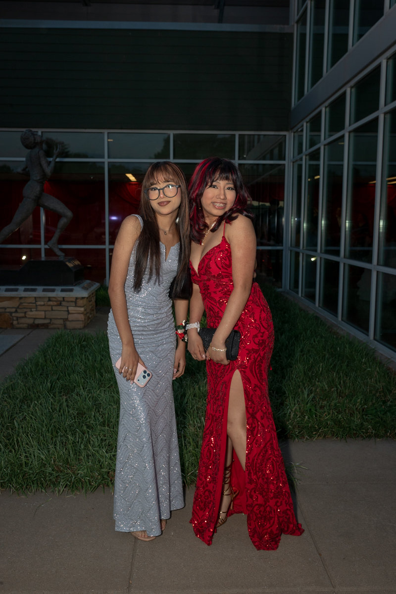 Two women in formal gowns pose outside a glass-front building at night, one in a silver dress holding a phone and the other in a red sequined dress with a slit.