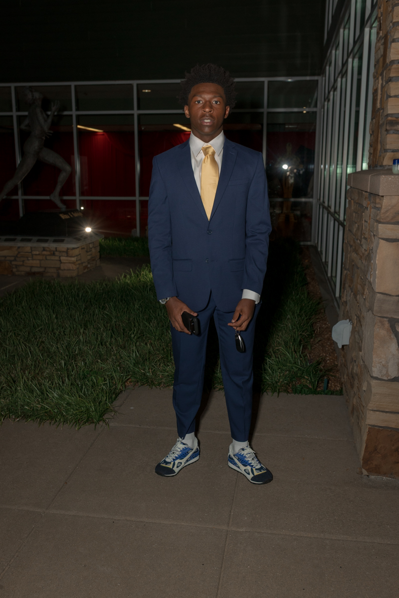 Young man in a navy suit with a yellow tie standing outdoors at night, holding sunglasses and a phone.