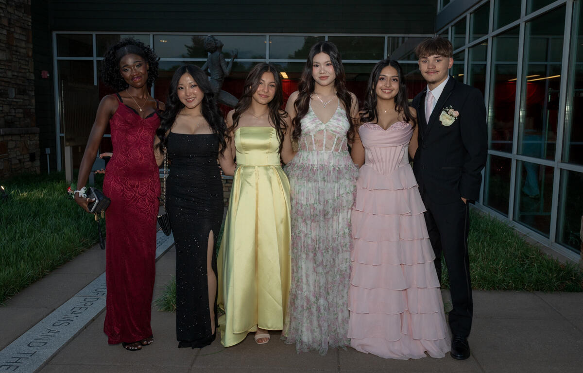 Six teenagers in formal attire pose together outdoors in front of a modern glass building, wearing colorful prom dresses and a dark suit with a boutonniere, smiling for the camera.