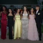 Six teenagers in formal attire pose together outdoors in front of a modern glass building, wearing colorful prom dresses and a dark suit with a boutonniere, smiling for the camera.