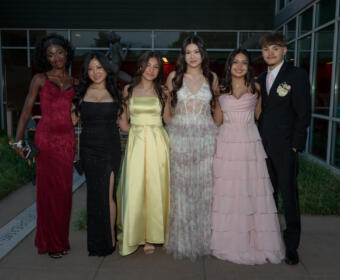 Six teenagers in formal attire pose together outdoors in front of a modern glass building, wearing colorful prom dresses and a dark suit with a boutonniere, smiling for the camera.