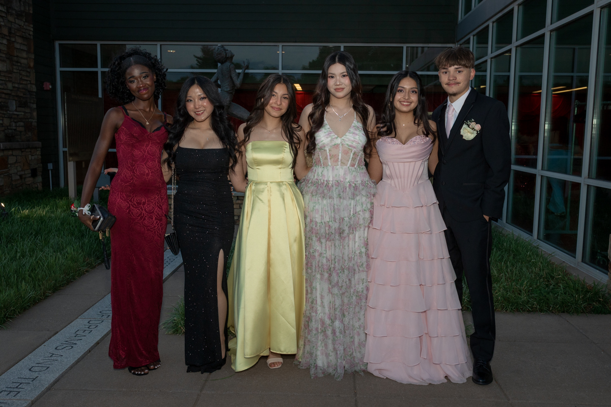 Six teenagers in formal attire pose together outdoors in front of a modern glass building, wearing colorful prom dresses and a dark suit with a boutonniere, smiling for the camera.