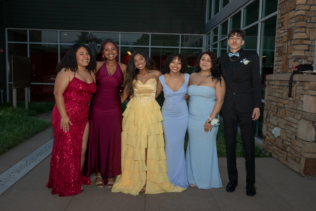Six teenagers in formal wear pose outside a modern building: red sequin gown, burgundy dress, yellow ruffled gown, and two light-blue gowns, with a young man in a black suit and bow tie nearby.