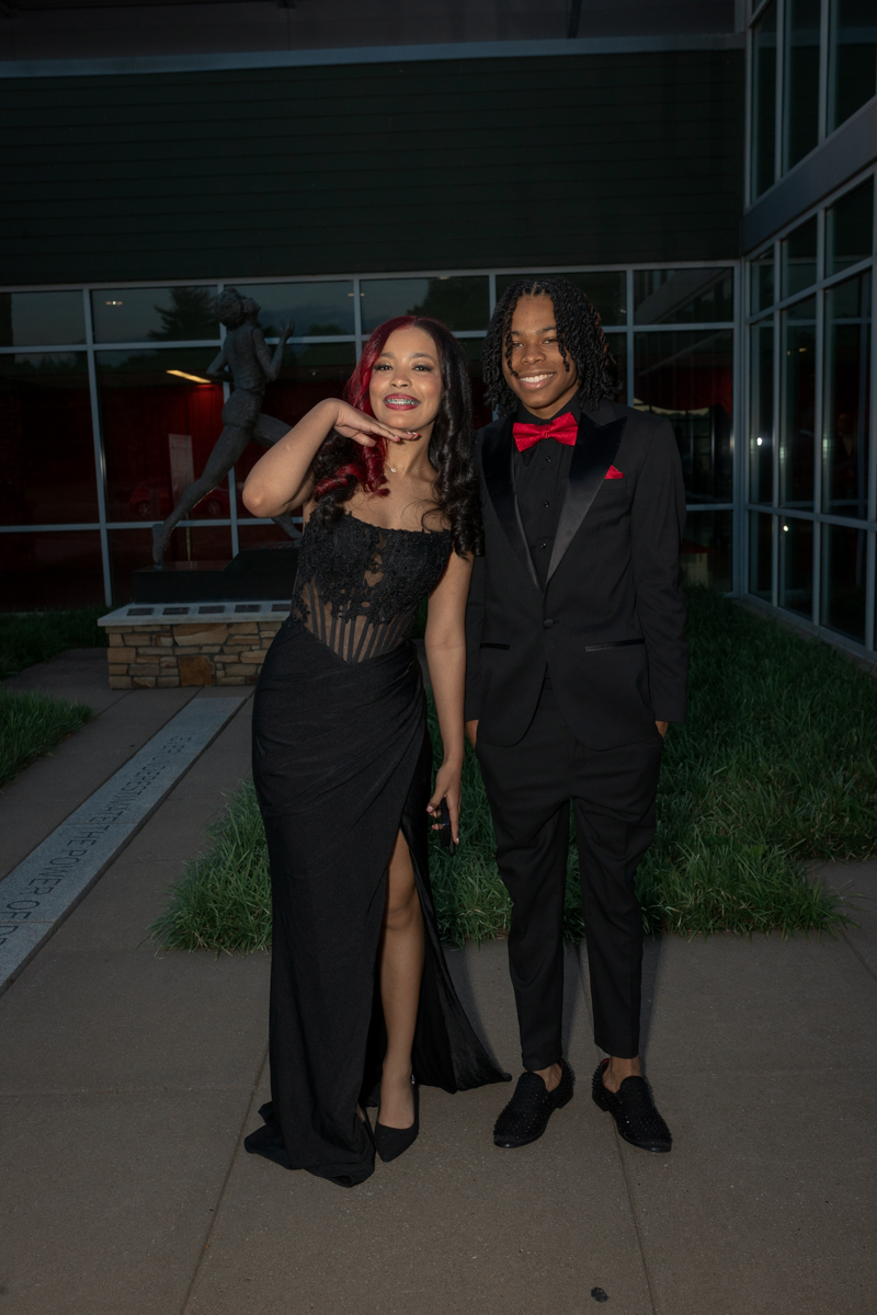 Two young adults in formal wear pose together outdoors at night, smiling at the camera outside a modern building.