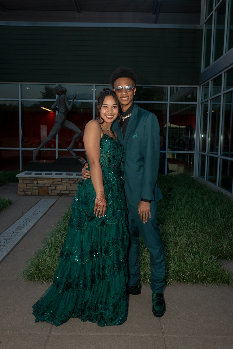 Couple posing outdoors in formal attire: woman in an emerald green sequined gown with henna on her hands and the man in a teal suit with sunglasses, standing beside a statue and glass building.