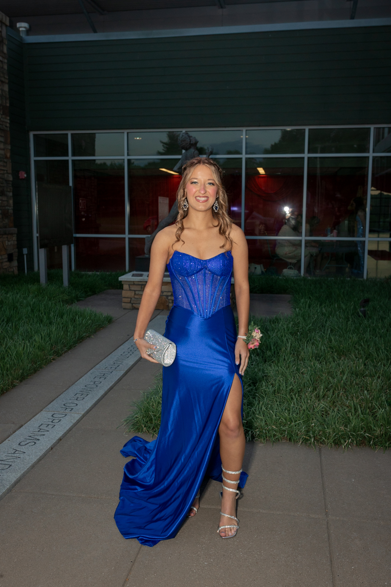 Young woman in a royal blue strapless gown with a corset bodice and thigh-high slit, posing outdoors with a silver clutch and floral boutonniere.