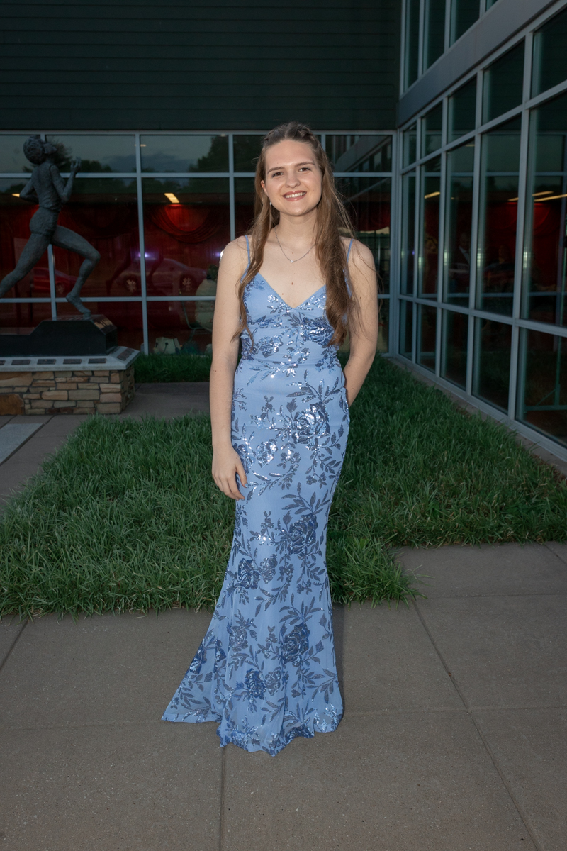 Young woman in a blue floral gown standing on a sidewalk outside a glass-front building, smiling at the camera.