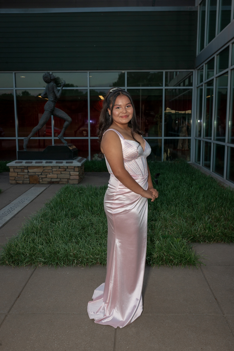 Young woman in a pale pink satin evening gown posing outdoors near a glass-front building and a statue in the background.