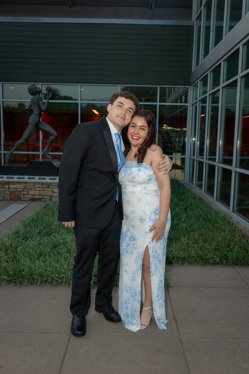 Couple in formal attire posing outside a glass-fronted building; man in a black suit with blue tie, woman in a light blue gown with a slit.
