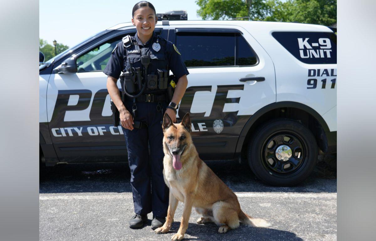 Female police officer in uniform standing beside a German Shepherd in front of a police SUV.