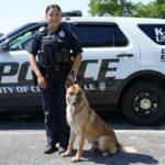 Female police officer in uniform standing beside a German Shepherd in front of a police SUV.
