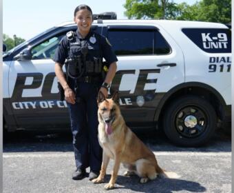 Female police officer in uniform standing beside a German Shepherd in front of a police SUV.