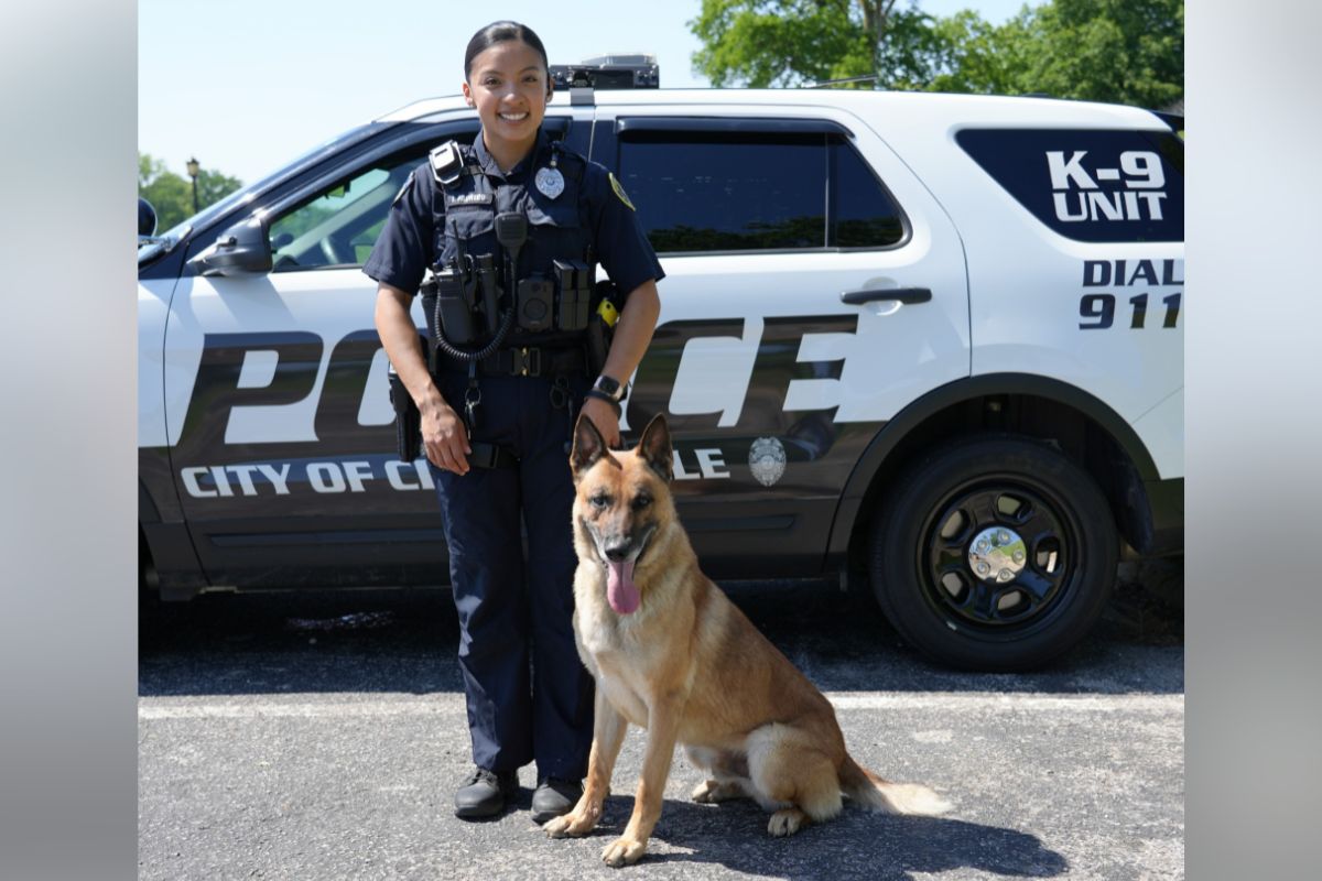 Female police officer in uniform standing beside a German Shepherd in front of a police SUV.