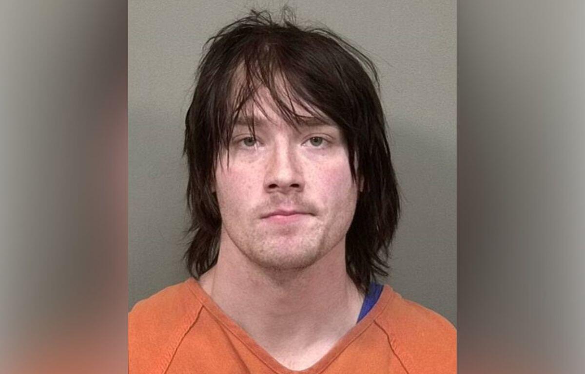 Mugshot of a man with shoulder-length dark hair in an orange prison jumpsuit against a gray background.