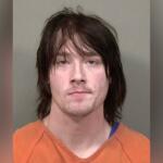 Mugshot of a man with shoulder-length dark hair in an orange prison jumpsuit against a gray background.