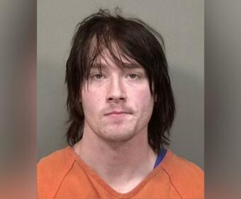 Mugshot of a man with shoulder-length dark hair in an orange prison jumpsuit against a gray background.