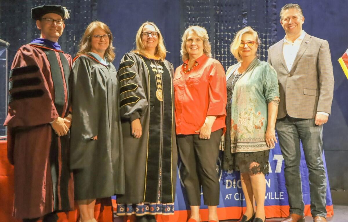 Group of seven adults on stage posing for a photo at a graduation event, some in academic gowns and robes.