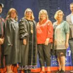 Group of seven adults on stage posing for a photo at a graduation event, some in academic gowns and robes.