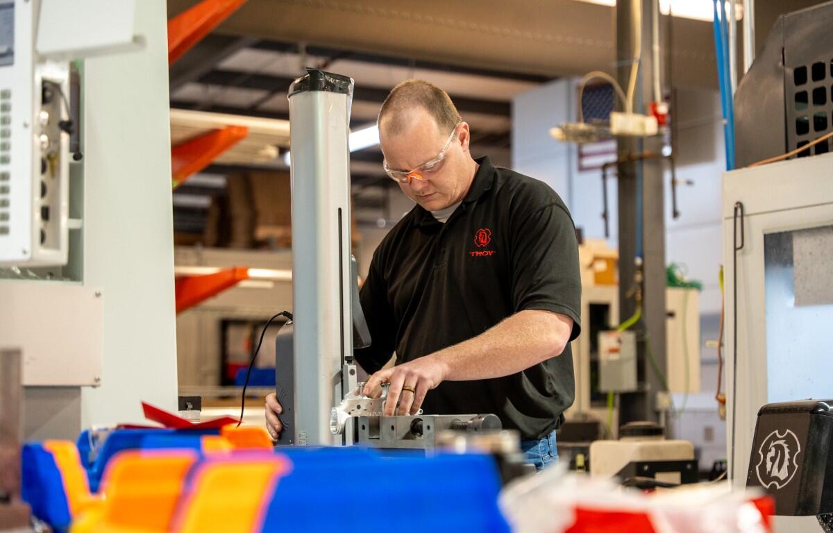 Machinist wearing safety glasses operates a milling machine in a busy workshop, with colorful bins in the foreground.