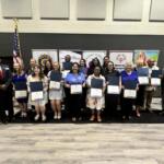 Diverse group on stage holding certificates during an awards ceremony, with banners and the American flag in the background.