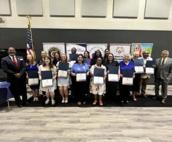 Diverse group on stage holding certificates during an awards ceremony, with banners and the American flag in the background.